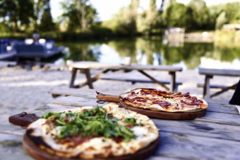 Deux pizzas sur une table en bois en plein air au bord d’un lac à Huttopia Vallée du Lot, parc de vacances en Nouvelle-Aquitaine, France.