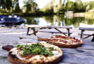 Deux pizzas sur une table en bois en plein air au bord d’un lac à Huttopia Vallée du Lot, parc de vacances en Nouvelle-Aquitaine, France.