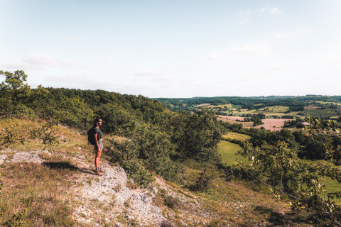 A hiker with a backpack stands overlooking lush hills and farmland at Huttopia Vallée du Lot in France.