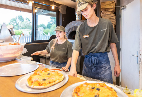 Twee jonge medewerkers maken pizza’s naast een houtoven op Huttopia Vallée du Lot, Nouvelle-Aquitaine.