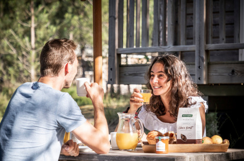 Pareja disfruta desayuno al aire libre con jugo y pan en Huttopia Vallée du Lot, Nouvelle-Aquitaine, Francia.