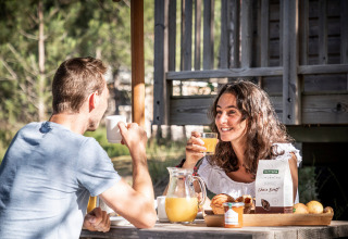 Pareja disfruta desayuno al aire libre con jugo y pan en Huttopia Vallée du Lot, Nouvelle-Aquitaine, Francia.