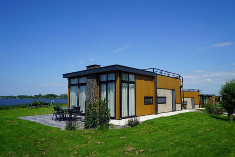 Lodge pavillon moderne au bord du lac, avec grandes baies vitrées, terrasse aménagée et pelouse verte.