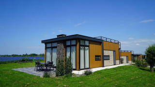 Moderna cabaña tipo pabellón junto al lago con grandes ventanales y terraza amueblada sobre césped verde.