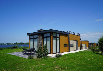 Moderne pavillon-lodge ved sø med store vinduer, terrasse og møbler udendørs på grøn græsplæne.