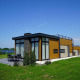 Moderne pavillon-lodge ved sø med store vinduer, terrasse og møbler udendørs på grøn græsplæne.