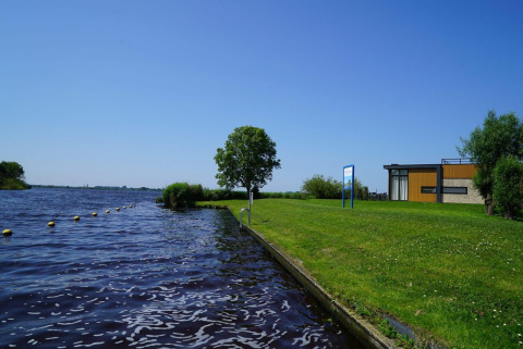 Foto del Pavilion en Kagerplassen, Países Bajos, con césped, agua, un árbol y edificio moderno al fondo.