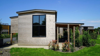 Tiny house moderna in legno con ampie finestre e giardino verde sotto un cielo azzurro limpido.