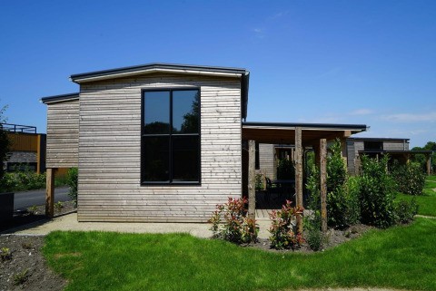 Tiny house moderna in legno con ampie finestre e giardino verde sotto un cielo azzurro limpido.