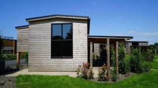 Tiny house moderna de madera con grandes ventanales y jardín verde bajo un cielo azul despejado.