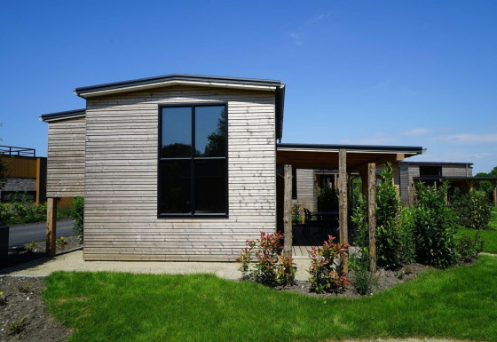Tiny house moderna de madera con grandes ventanales y jardín verde bajo un cielo azul despejado.