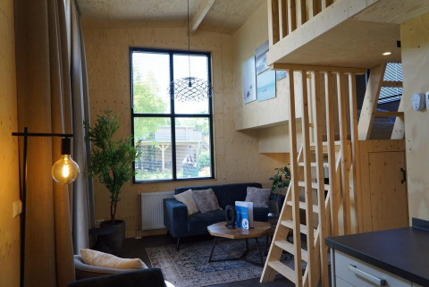Interno di Tiny House vicino a Kagerplassen, nei Paesi Bassi, con soppalco in legno, divano e grandi finestre.
