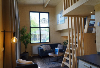 Vista interior de Tiny House en Kagerplassen, Países Bajos, con sofá, altillo de madera y ventana grande.