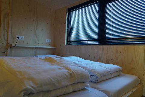 Camera da letto accogliente in una tiny house ai Kagerplassen, Paesi Bassi, con letto e grandi finestre a veneziana.