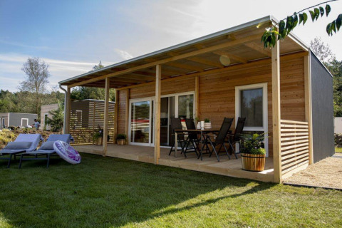 Moderne houten lodge met overdekt terras, tuinmeubels, ligstoelen en groene graspartij in zon.