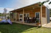 Modern wooden lodge with covered terrace, outdoor dining set, lounge chairs, and lush green lawn.