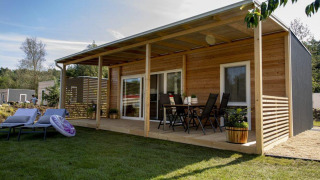 Modern wooden lodge with covered terrace, outdoor dining set, lounge chairs, and lush green lawn.