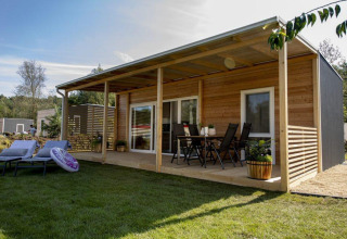 Moderne houten lodge met overdekt terras, tuinmeubels, ligstoelen en groene graspartij in zon.