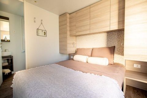 Lichte lodge-slaapkamer met tweepersoonsbed, houten kasten, kussens en zicht op badkamer in de hoek.