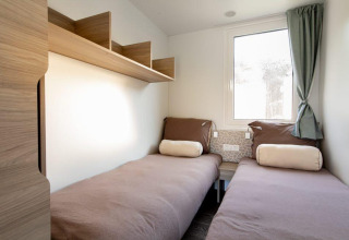 Lichte lodgekamer met twee eenpersoonsbedden, bruine dekens, houten planken en raam met gordijn.