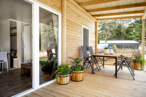 Gezellige lodge met houten terras, tuinmeubilair en plantenbakken, omgeven door bos en zonlicht.