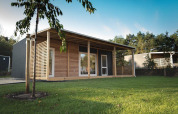Moderna cabaña con fachada de madera y gran terraza, rodeada de césped verde y árboles frondosos.