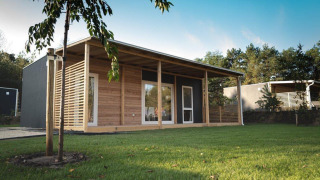 Moderna cabaña con fachada de madera y gran terraza, rodeada de césped verde y árboles frondosos.