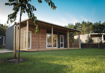 Modern lodge with wooden exterior and large veranda, set on lush green grass and surrounded by trees.