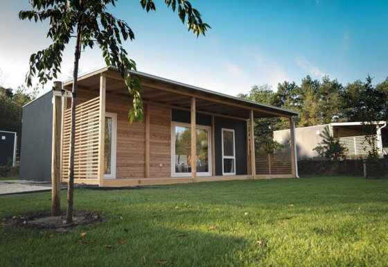 Moderne lodge met houten gevel en ruime veranda, omgeven door grasveld en bomen onder blauwe lucht.