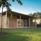 Moderne lodge met houten gevel en ruime veranda, omgeven door grasveld en bomen onder blauwe lucht.
