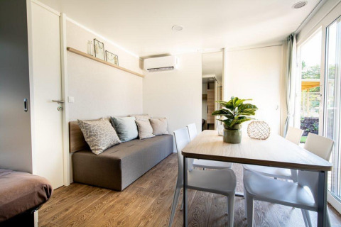 Lichte lodge met grijze zetel, eettafel, witte stoelen, kamerplant en grote ramen naar buiten toe.