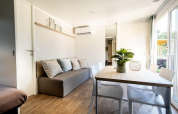 Acogedora sala de lodge con sofá gris, mesa de comedor, sillas blancas, planta y grandes ventanas.