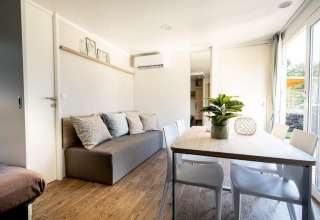 Acogedora sala de lodge con sofá gris, mesa de comedor, sillas blancas, planta y grandes ventanas.