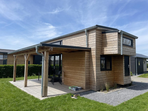 Acogedora Tiny House Plus de madera con terraza cubierta, muebles de exterior y césped verde alrededor.
