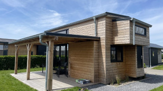 Acogedora Tiny House Plus de madera con terraza cubierta, muebles de exterior y césped verde alrededor.