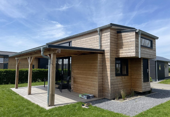 Moderne Tiny House Plus lodge van hout met overdekt terras, tuinmeubilair en groen gazon buiten.