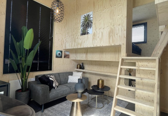 Moderne stue i Tiny House Plus-lodge i De Rijp, Holland, med trappe, sofa og store vinduer.