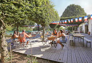 Udendørs terrasse på Huttopia Royat i Auvergne-Rhône-Alpes med gæster, træer og farverige guirlander.