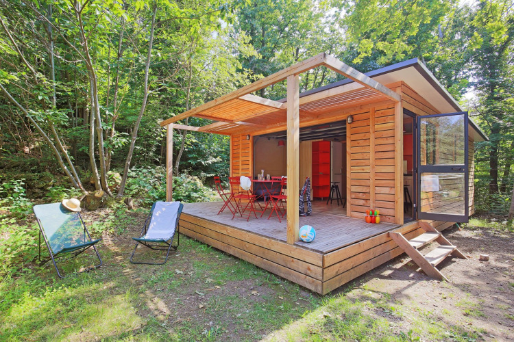 Holzhütte mit Terrasse und Gartenstühlen im Wald bei Huttopia Royat, Auvergne-Rhône-Alpes, Frankreich.