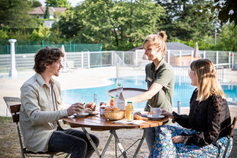 Un couple prend le petit-déjeuner en plein air près de la piscine au Huttopia Royat en Auvergne-Rhône-Alpes.