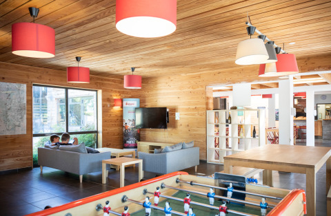 Sala común moderna en Huttopia Royat, Francia, con sofás, futbolín, techos de madera y lámparas rojas.