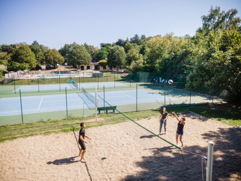 Holiday park scene with people playing beach volleyball, tennis courts, and a pool at Huttopia Royat, France.