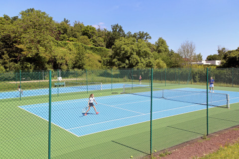 Børn og voksne spiller tennis på udendørsbaner omgivet af grønne træer i Huttopia Royat, Frankrig.