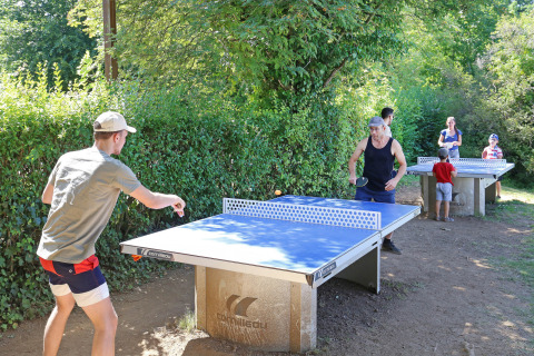 Personas jugando al tenis de mesa al aire libre en Huttopia Royat, un parque vacacional en Auvergne-Rhône-Alpes, Francia, rodeados de vegetación.