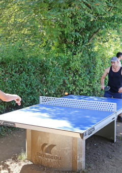 Personas jugando al tenis de mesa al aire libre en Huttopia Royat, un parque vacacional en Auvergne-Rhône-Alpes, Francia, rodeados de vegetación.
