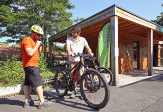 Twee mannen met fietsen voor een houten chalet in Huttopia Royat, vakantiepark in Auvergne-Rhône-Alpes.