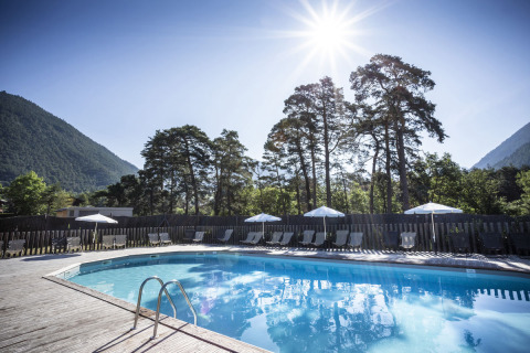 Piscina al aire libre en Huttopia Bourg Saint-Maurice, con montañas y árboles al fondo en un día soleado.