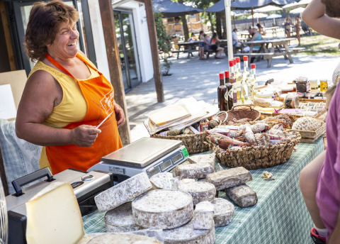 Mujer sonriente con delantal naranja vende quesos y embutidos en un mercado al aire libre, Huttopia Bourg Saint-Maurice, Francia.