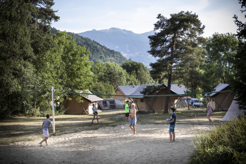 Personas juegan al voleibol en una cancha de arena en un camping rodeado de cabañas, árboles y montañas.