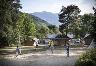 Vakantiegangers spelen volleybal op een zandveld in een vakantiepark met hutten en bergen op de achtergrond.